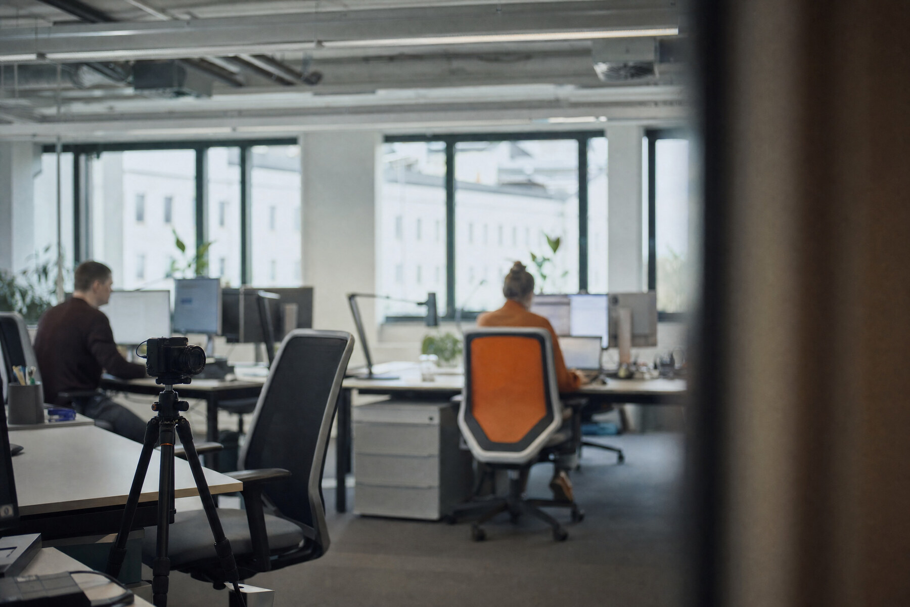 Kamera auf einem Stativ richtet sich auf leere Schreibtische in einem Großraumbüro, Beschäftigte arbeiten im Hintergrund
