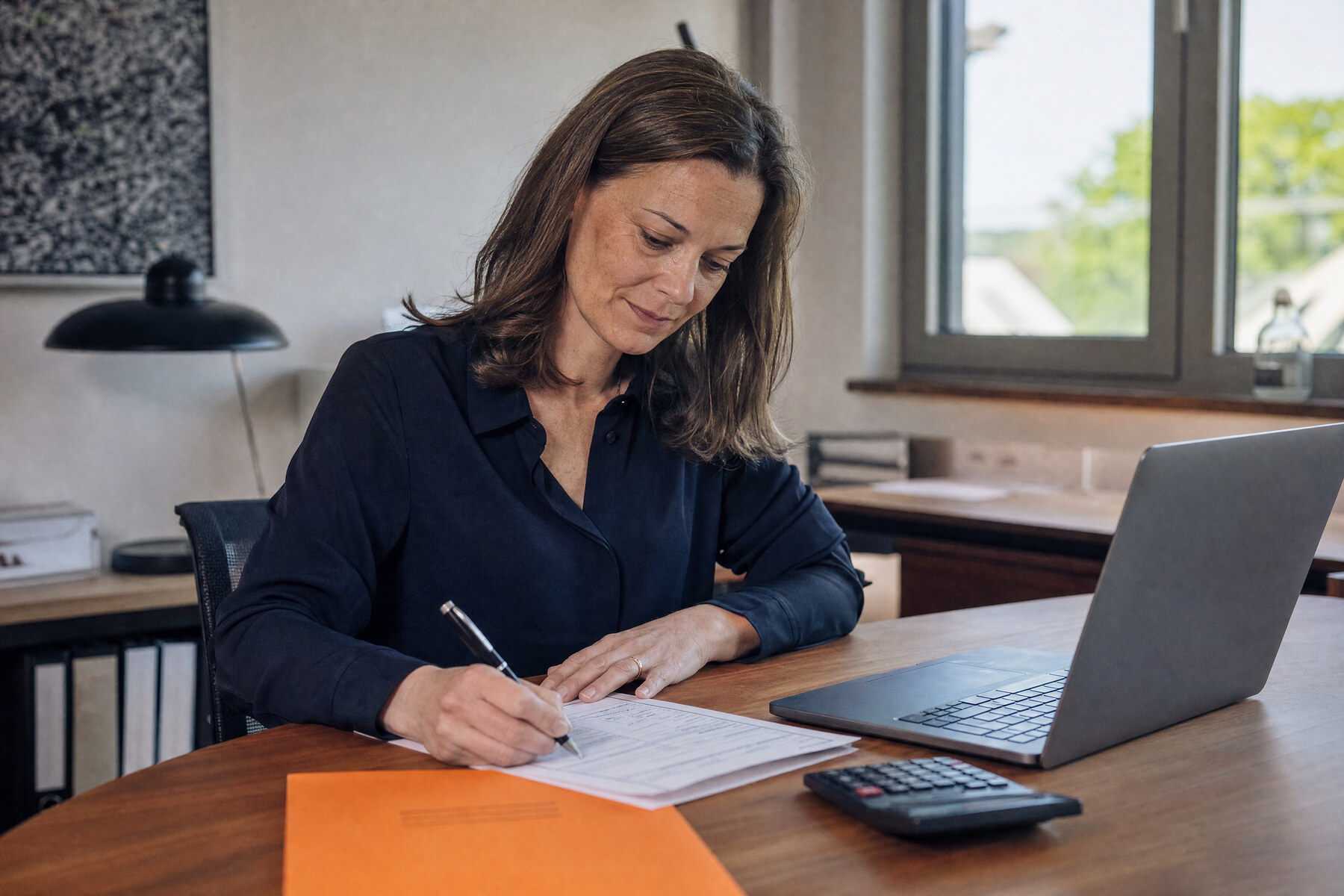 Geschäftsführerin füllt am Schreibtisch einen Förderantrag aus, daneben Laptop, Taschenrechner und orangefarbene Mappe