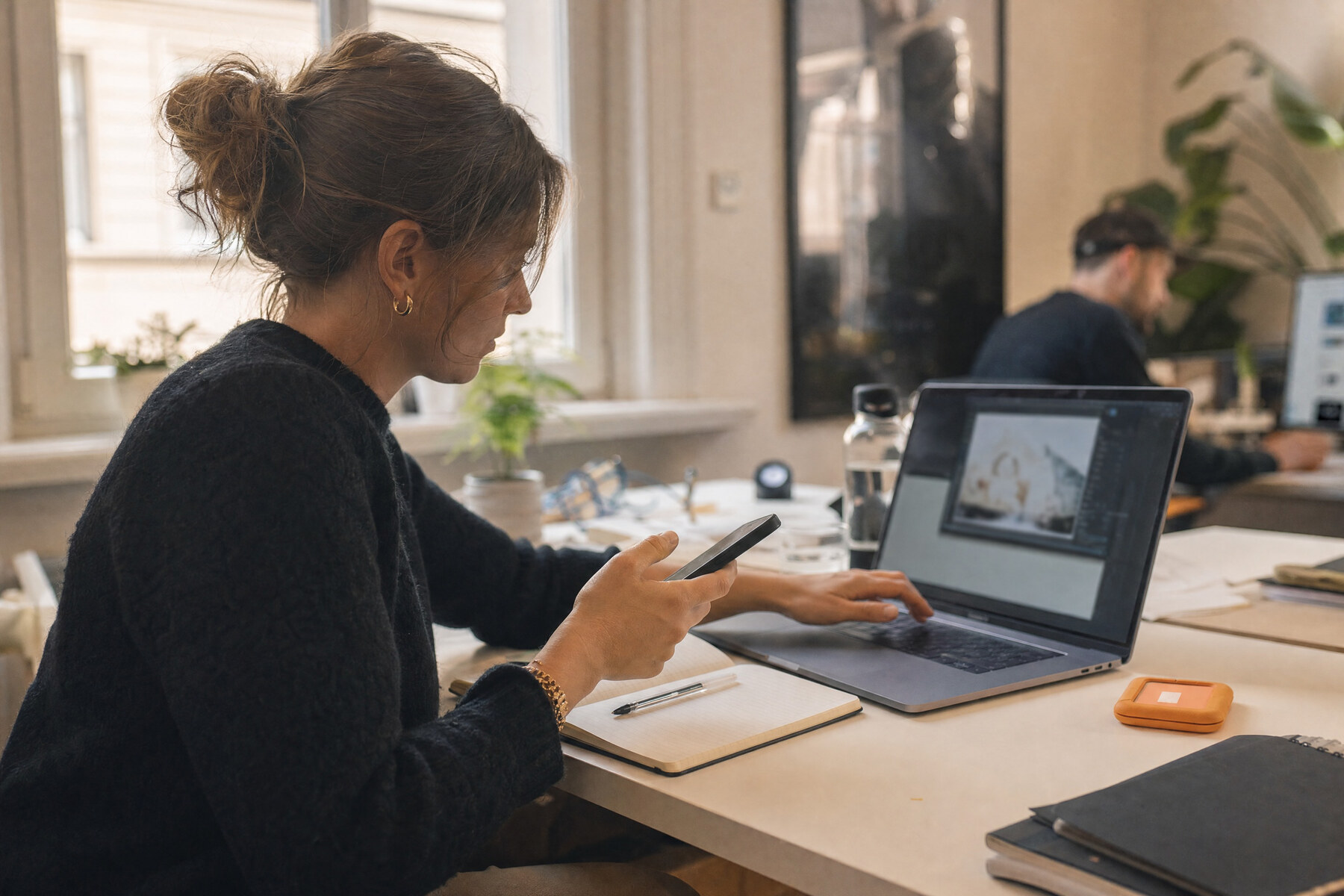 Social-Media-Managerin mit Smartphone und Laptop-Bildbearbeitung am Schreibtisch, daneben ein orangefarbenes Etikett