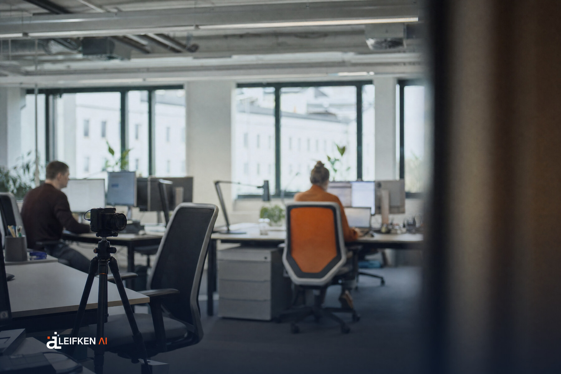 Kamera auf einem Stativ richtet sich auf leere Schreibtische in einem Großraumbüro, Beschäftigte arbeiten im Hintergrund
