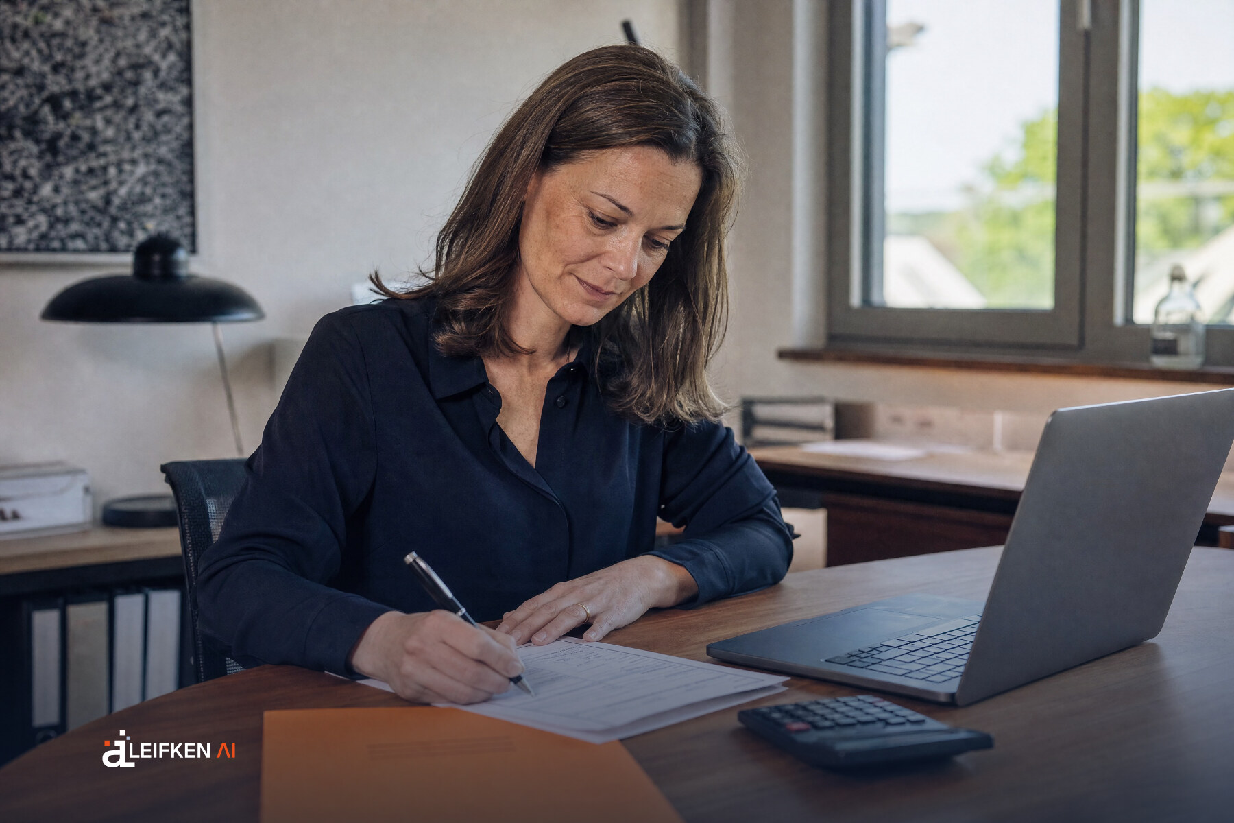 Geschäftsführerin füllt am Schreibtisch einen Förderantrag aus, daneben Laptop, Taschenrechner und orangefarbene Mappe
