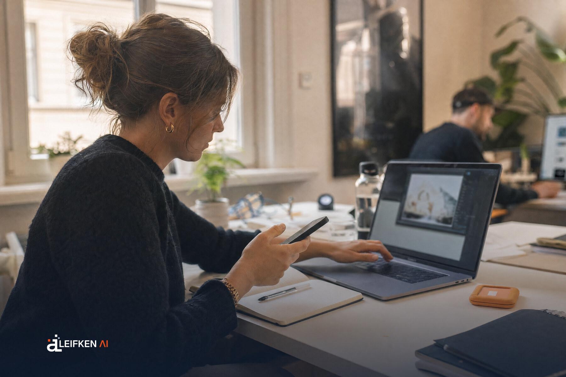 Social-Media-Managerin mit Smartphone und Laptop-Bildbearbeitung am Schreibtisch, daneben ein orangefarbenes Etikett